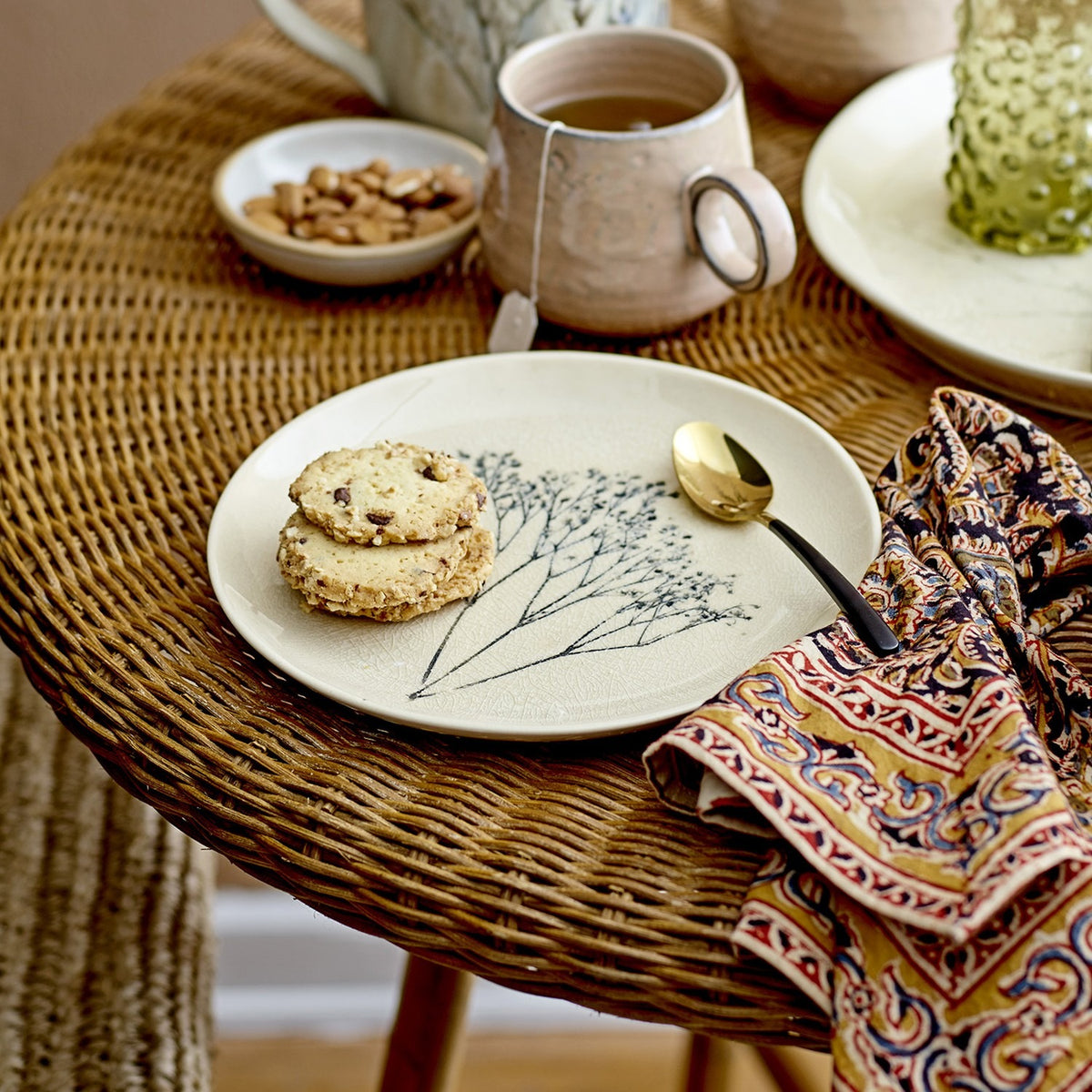 Cream crackle stoneware side plate from the Bea dinner set, with botanical grass motif, shown on a wicker table with cookies, a gold spoon, and a tea mug.