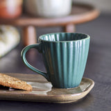 Latina Stoneware Cup 220ml in Green with fluted ribbed sides, glossy glaze and open loop handle, shown on a speckled plate beside an oat biscuit.