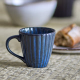 Latina Stoneware Cup 220ml in blue with a fluted, ribbed body and looped handle, shown on a table beside a white bowl and pastry.