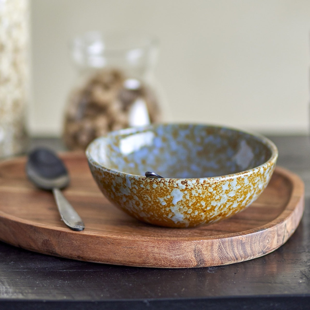 Paula Mottled Stoneware Bowl 16cm in brown and blue reactive glaze, shown on a wooden tray with a spoon and jar in a kitchen setting.