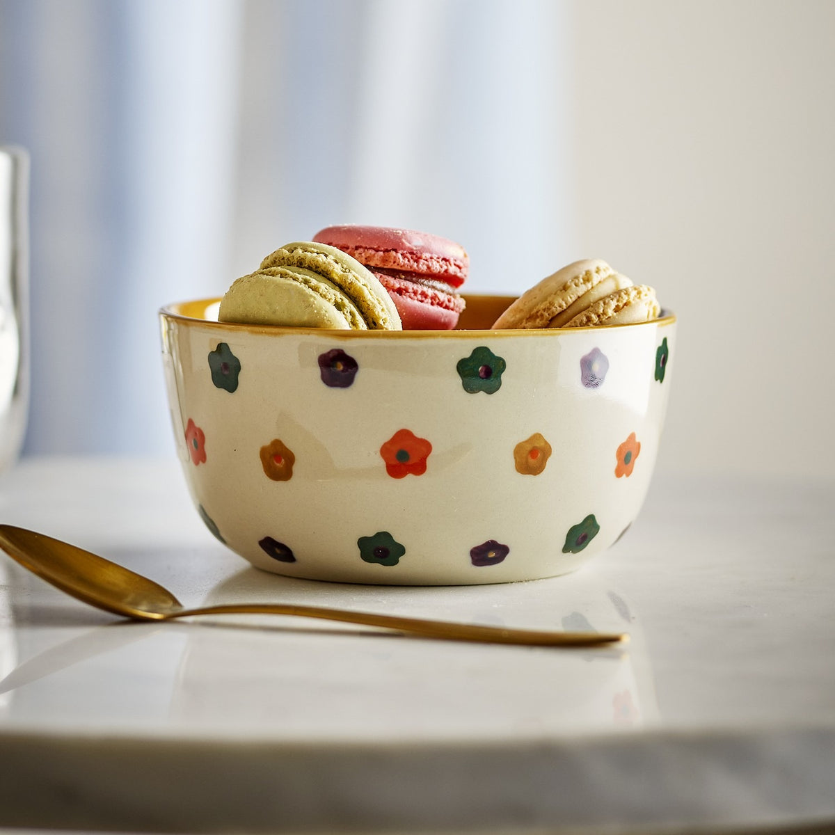Addy Stoneware Bowl 12cm in cream with hand-painted multicoloured flowers and a yellow-glazed rim, filled with macarons beside a gold spoon on a marble surface.