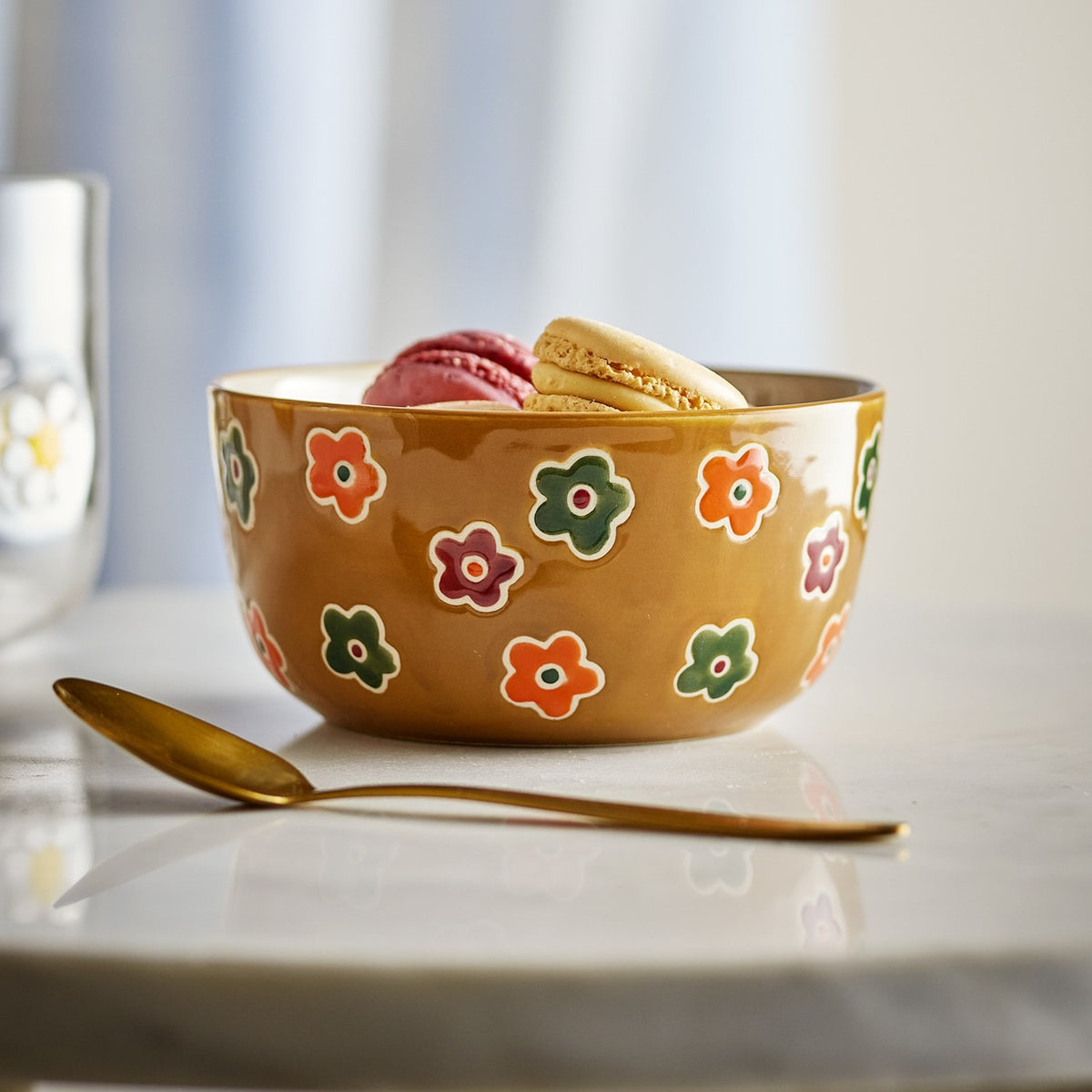 Addy Stoneware Bowl 12cm in Brown with hand-painted floral pattern, shown holding macarons beside a brass spoon on a marble tabletop.