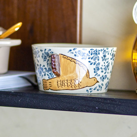 Hezha Floral Stoneware Bowl 13cm in cream, hand-painted with blue floral pattern and a raised bird in relief, shown on a dark shelf in a home setting.