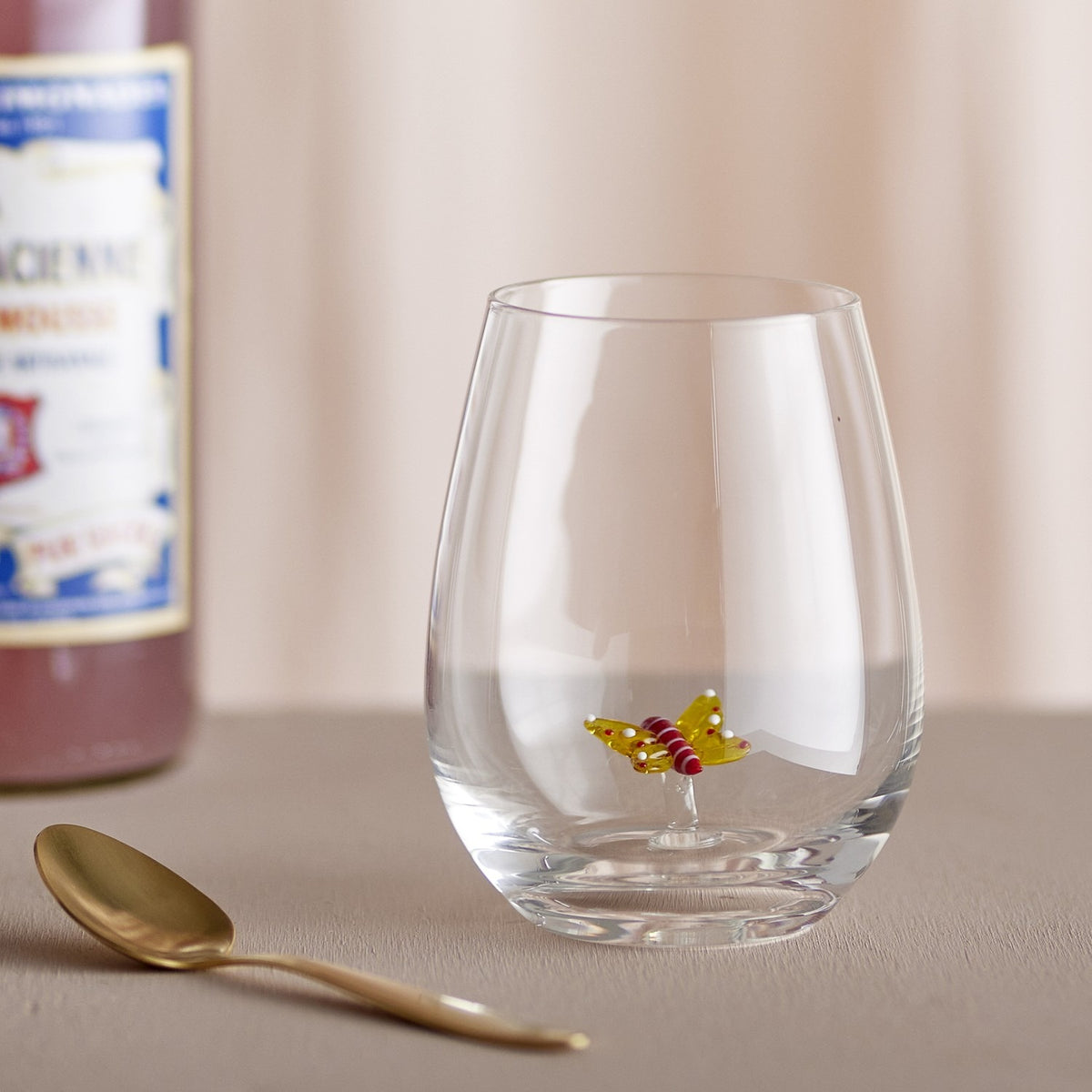 Misa Butterfly Tumbler in clear mouth-blown glass with a small decorative butterfly on a stalk inside the bowl, stemless rounded shape with a thick base, shown beside a gold spoon and bottle.