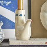 Trudy Bird Stoneware Candle Holder in cream, a dove-shaped ceramic piece with a painted wing and red-tipped beak, holding a lit taper candle on a shelf with books and artwork.