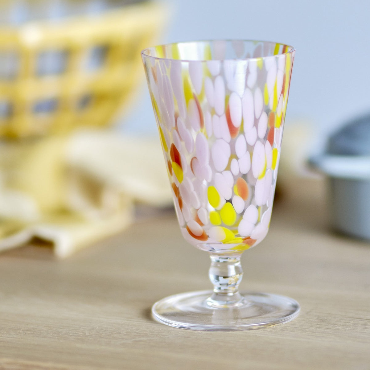 Lilya Speckled Wine Glass in Pink, flared glass bowl with pink, yellow and orange speckles, short stem and flat foot, shown on a wooden surface.