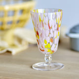 Lilya Speckled Wine Glass in Pink, flared glass bowl with pink, yellow and orange speckles, short stem and flat foot, shown on a wooden surface.