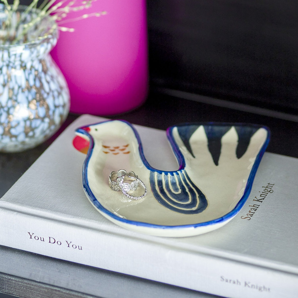 Trudy Bird Stoneware Bowl in white, a bird-shaped ceramic trinket dish with blue-striped tail and orange markings, shown holding a ring on a book beside a pink candle and patterned vase.