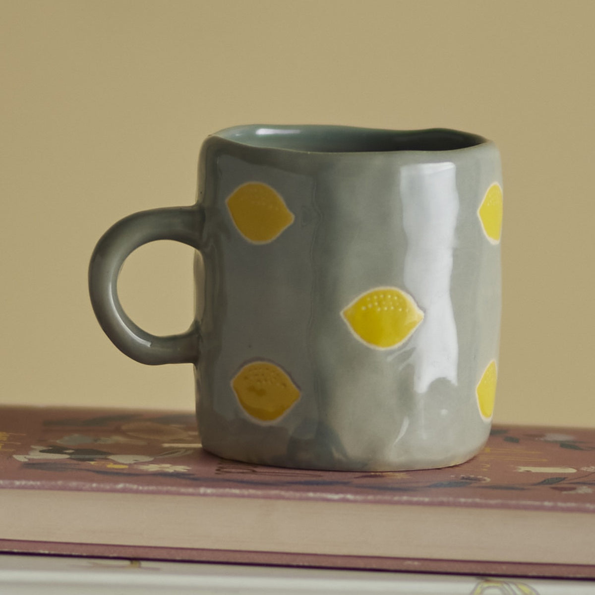 Agnes Stoneware Cup 230ml in green, a small ceramic mug with a curved handle and glossy finish, dotted with yellow lemon motifs, shown on a book against a plain beige background.