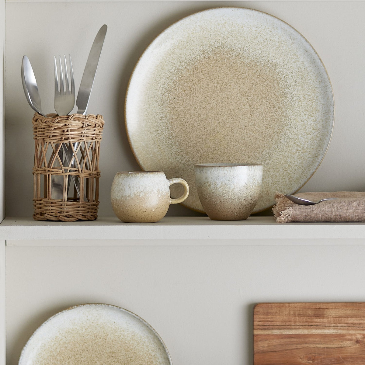 Paula Stoneware Breakfast Set of 12 in beige reactive glaze, showing a large plate, two cups with loop handles, cutlery in a woven holder, and a wooden board on a shelf.