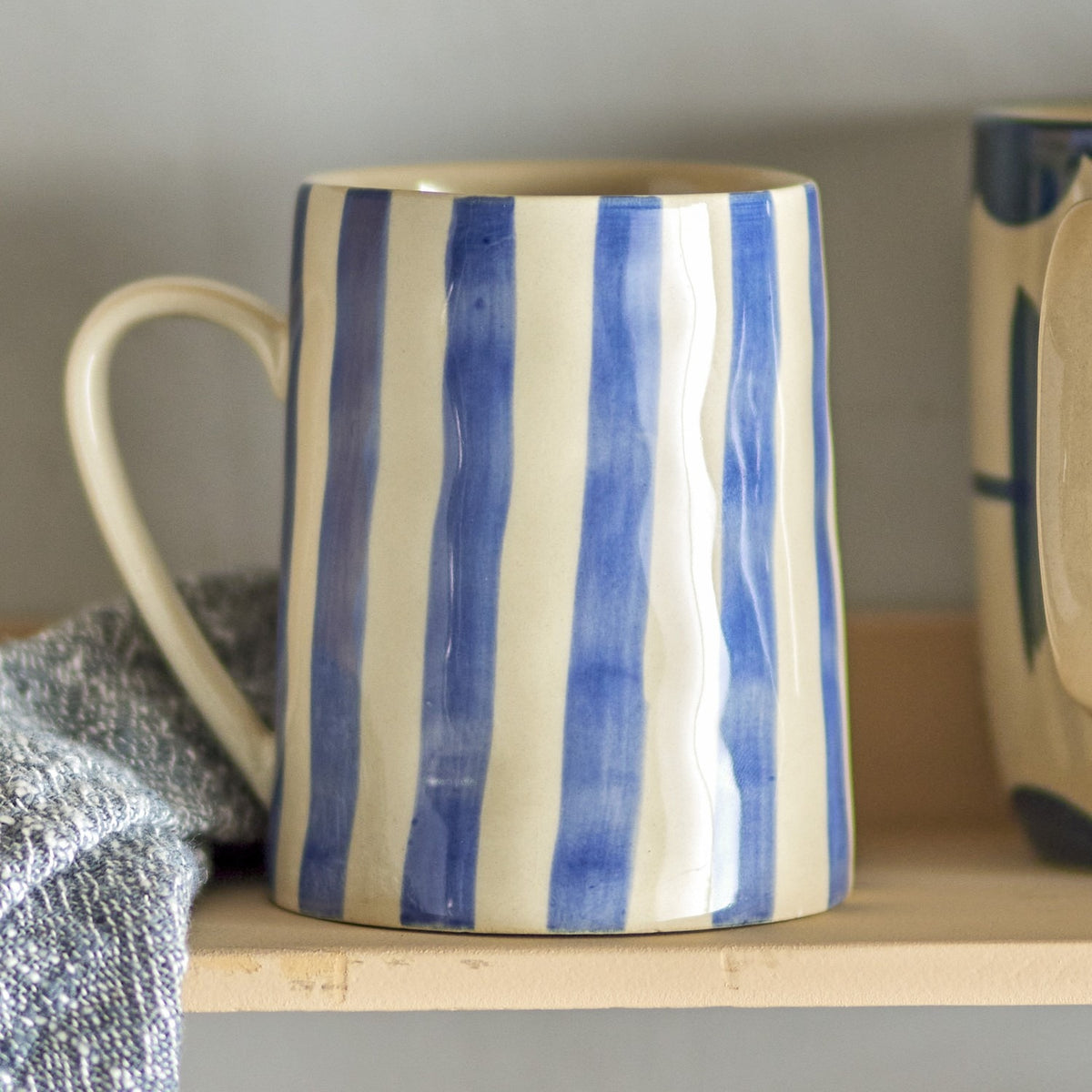 Begonia Striped Stoneware Mug 440ml in Blue, hand-painted vertical blue stripes with a loop handle, shown on a wooden shelf beside folded fabric.
