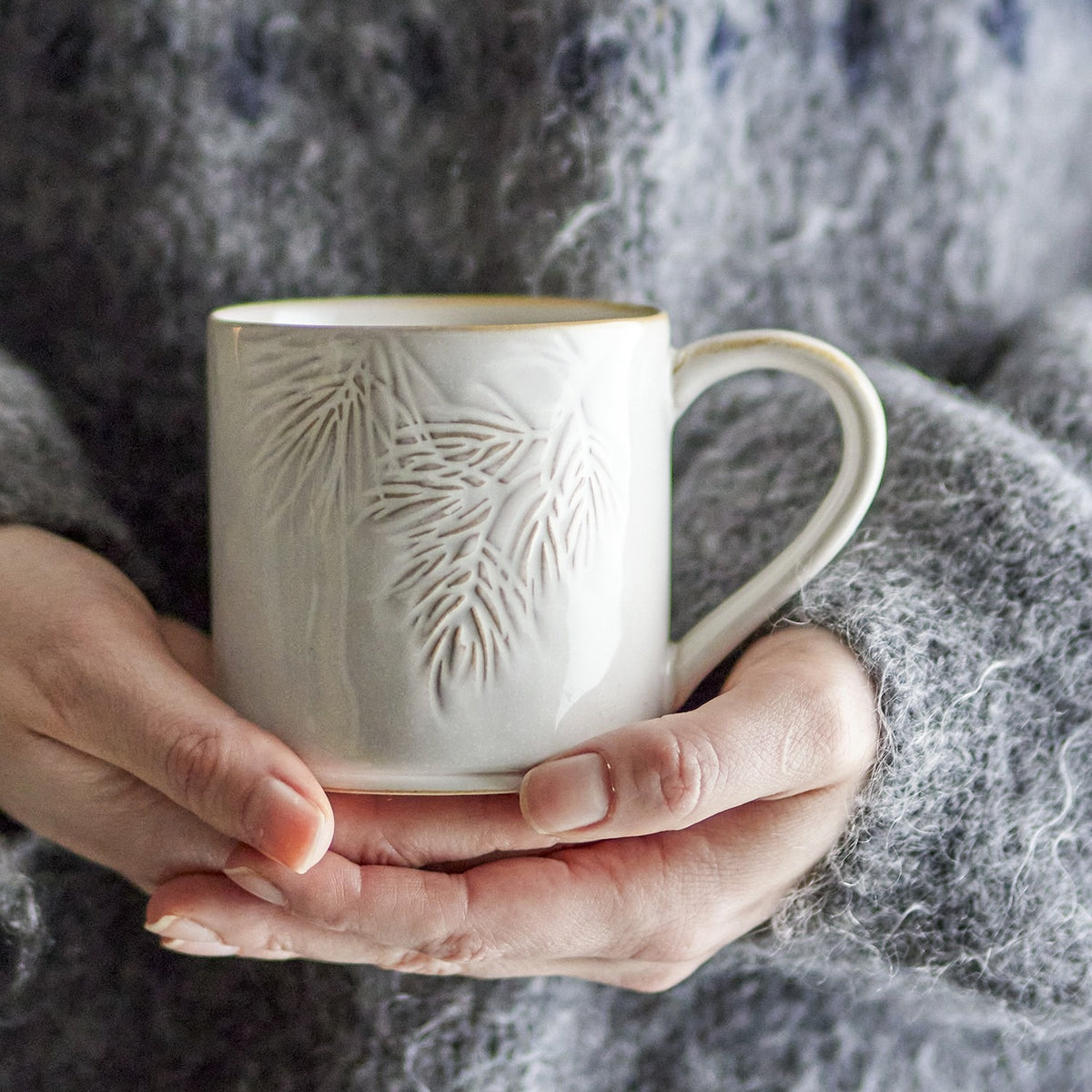 Hands cradle the Winter Embossed Stoneware Christmas Cup in cream, showing its pine needle relief and gold-tone rim against a soft grey knit background.