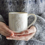 Hands cradle the Winter Embossed Stoneware Christmas Cup in cream, showing its pine needle relief and gold-tone rim against a soft grey knit background.