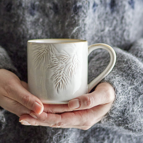 Hands cradle the Winter Embossed Stoneware Christmas Cup in cream, showing its pine needle relief and gold-tone rim against a soft grey knit background.
