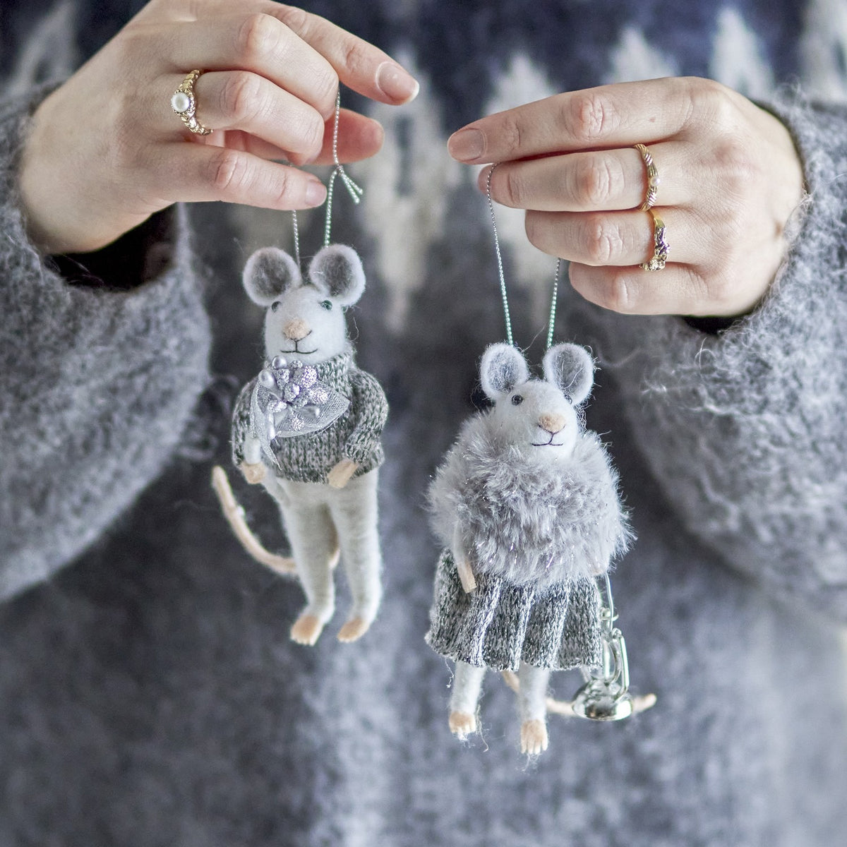Two hands holding the Peo Wool Christmas Tree Decorations, felted grey mice in a knitted jumper and fluffy stole, against a grey sweater background.