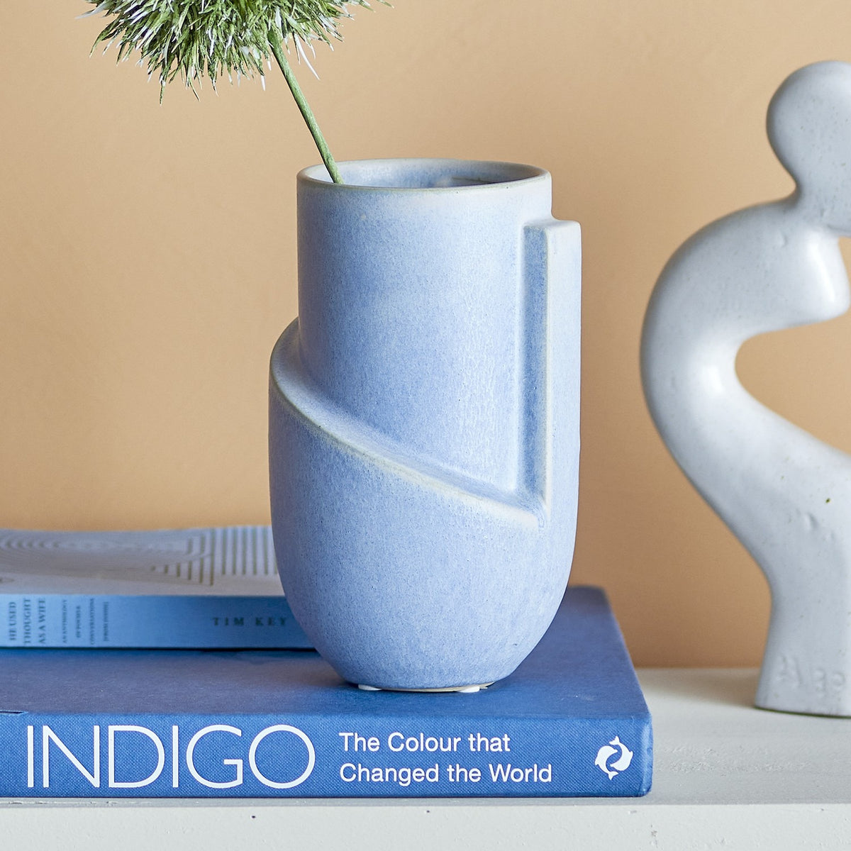 Norbert Stoneware Vase 18.5cm in mottled blue with sculpted diagonal band and ridge detail, shown holding a dried flower stem atop a stack of books beside a pale figurine sculpture.