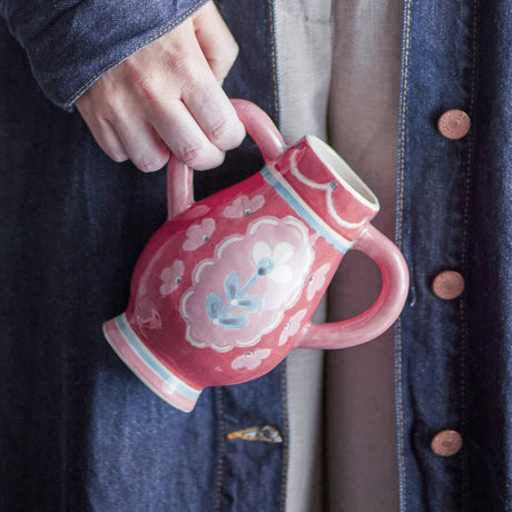 Odina Floral Stoneware Vase 15cm in red, held by hand, showing its rounded body, loop handles, and hand-painted floral motif against a denim jacket background.