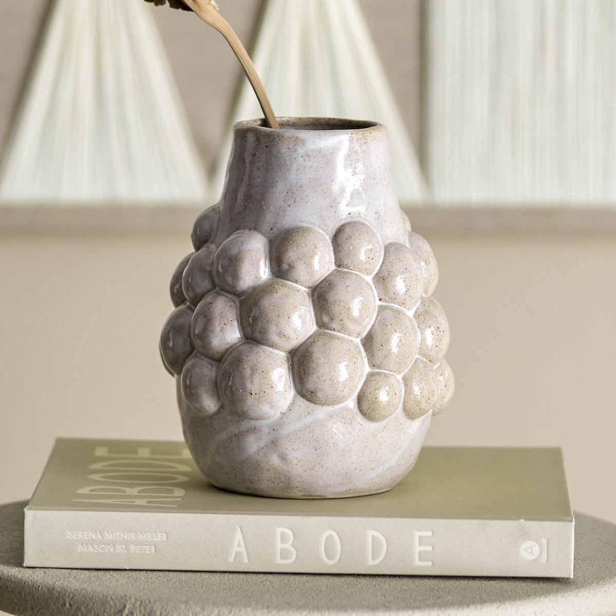 Poldor Stoneware Vase 19cm in cream with a cluster of raised bobbles around the middle and a narrow neck, shown holding a dried stem on a book in a living room setting.