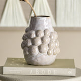 Poldor Stoneware Vase 19cm in cream with a cluster of raised bobbles around the middle and a narrow neck, shown holding a dried stem on a book in a living room setting.