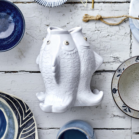 Fili Fish Stoneware Vase 19cm in white, shaped as two fish standing tail-to-tail with relief scale patterns, raised eyes and curled tail feet, shown among blue and white tableware.