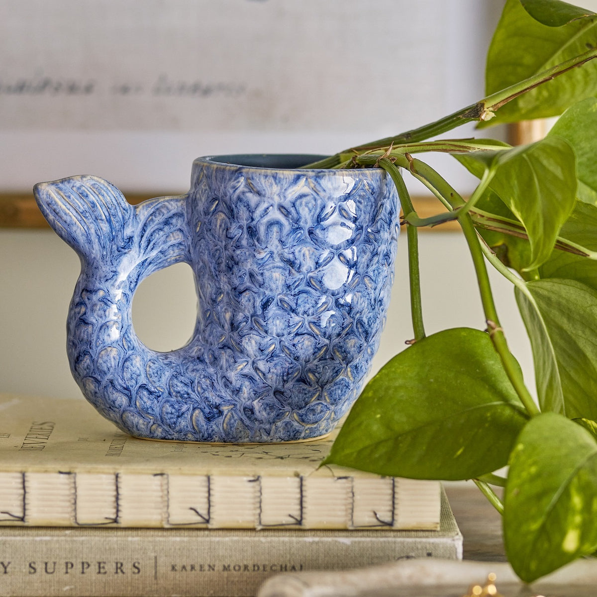Ariella Stoneware Vase 12cm in Blue, shaped like a curling fish tail with scale-textured body and a looped fin handle, displayed on books beside a trailing houseplant.