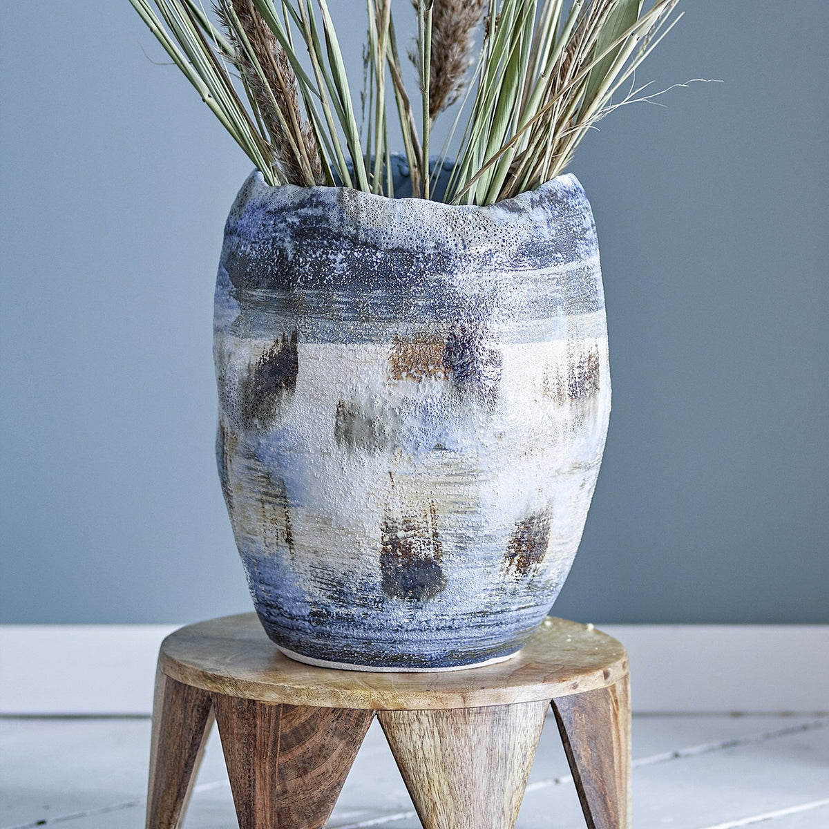 Nienna Stoneware Vase 31cm in Blue with a wavy rim and uneven glazed surface in blue, grey and tan tones, shown on a wooden stool with dried grasses.