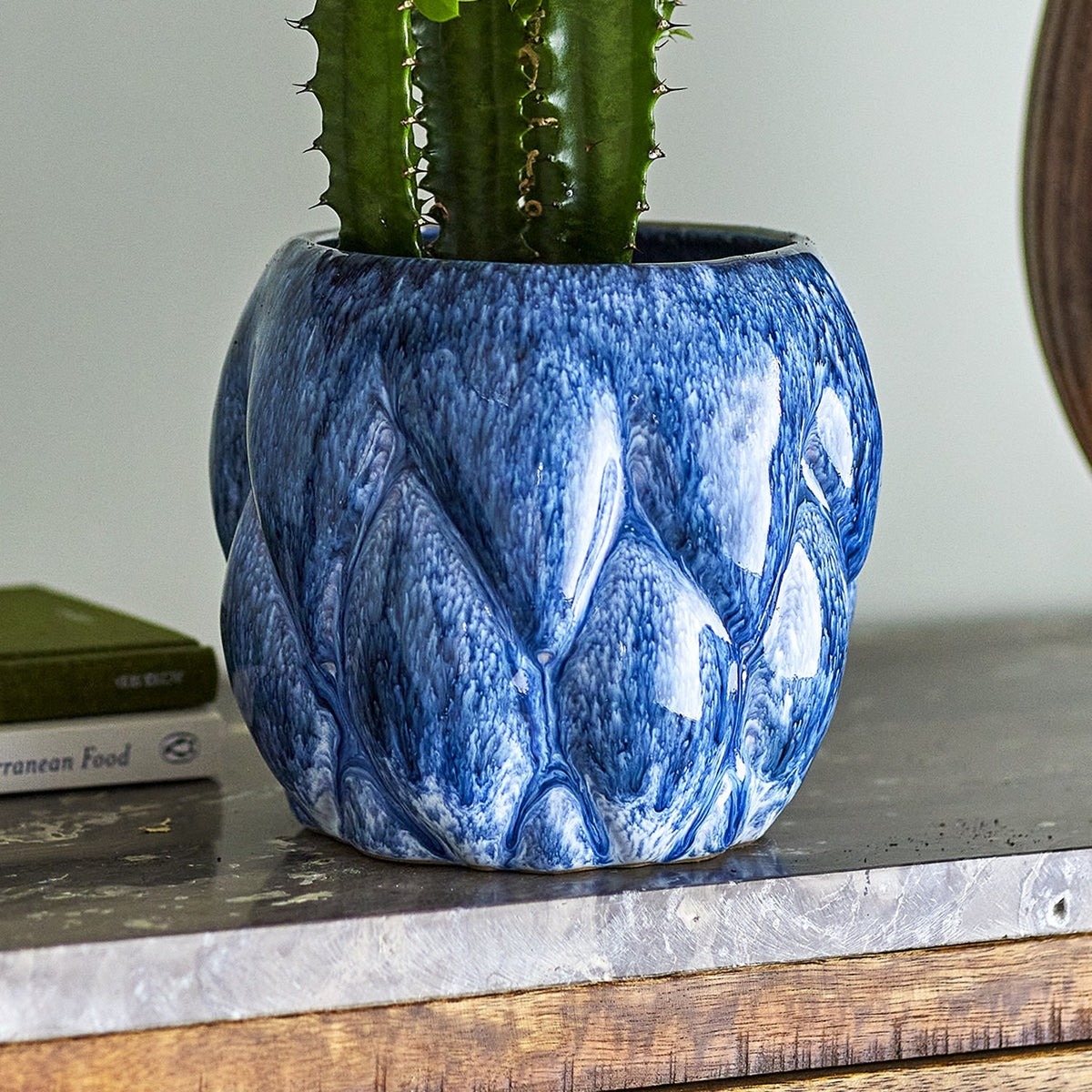 Posey Stoneware Plant Pot in Blue, a rounded ceramic planter with raised, overlapping pointed segments and a glossy blue glaze, shown holding a cactus on a wooden shelf.