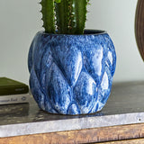 Posey Stoneware Plant Pot in Blue, a rounded ceramic planter with raised, overlapping pointed segments and a glossy blue glaze, shown holding a cactus on a wooden shelf.