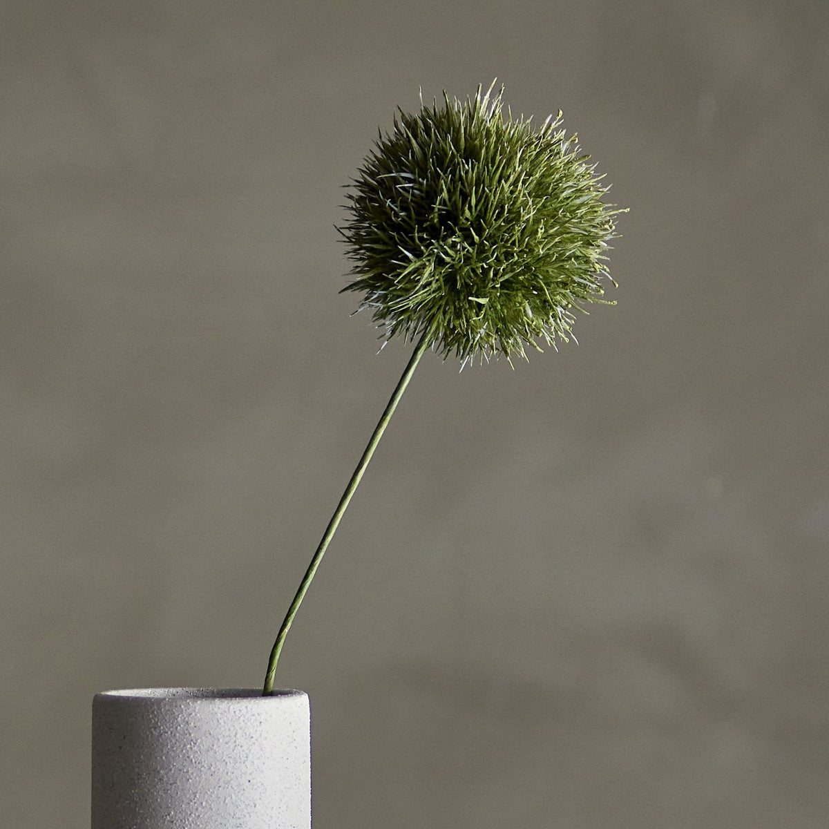 Artificial allium stem in green with a round, spiky globe-shaped head on a slender curved stem, displayed in a light grey cylindrical vase against a neutral background.