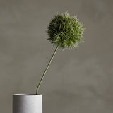 Artificial allium stem in green with a round, spiky globe-shaped head on a slender curved stem, displayed in a light grey cylindrical vase against a neutral background.