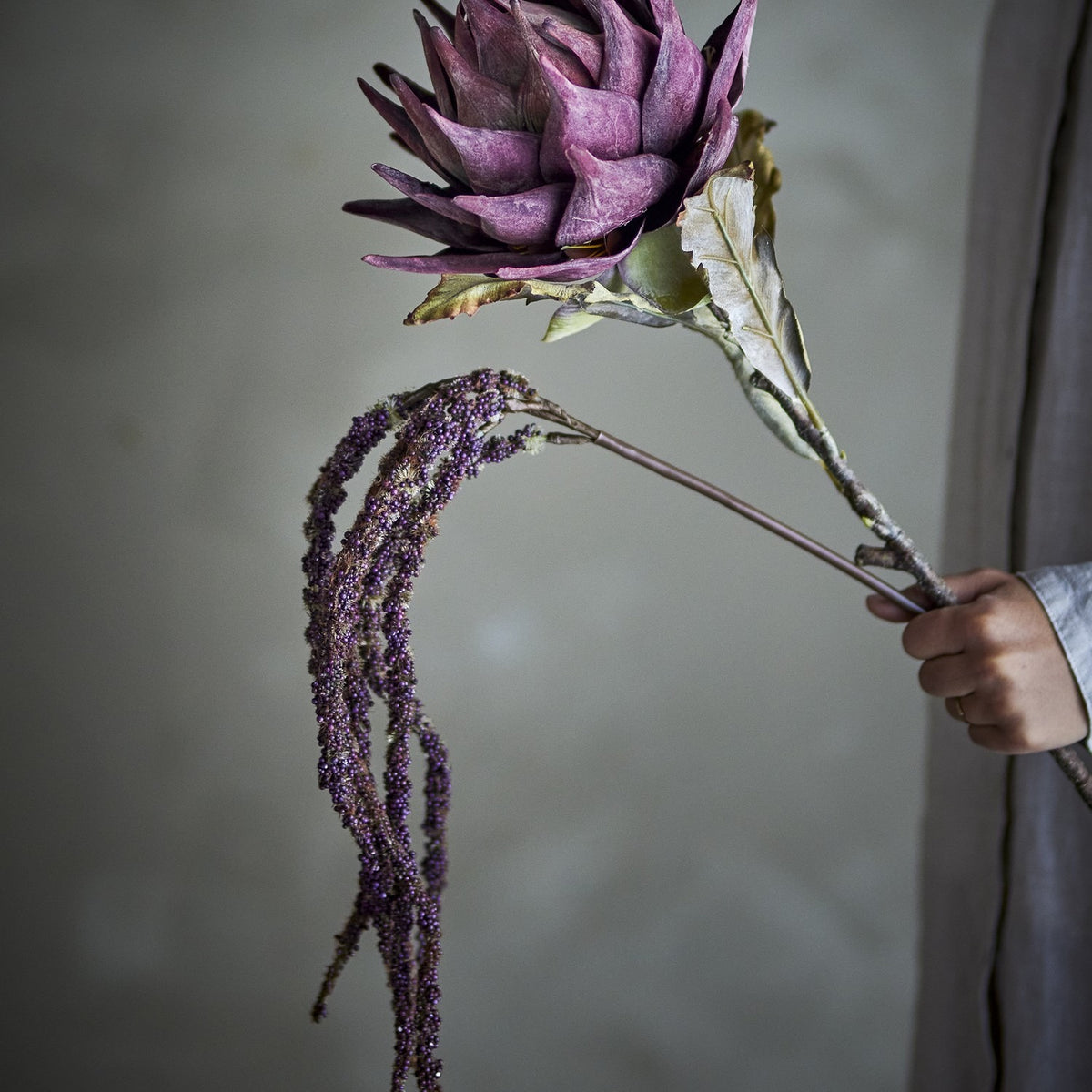 Protea Artificial Stem 76cm in purple, held by hand to show the layered petal flower head with trailing purple amaranthus-style strands, against a neutral indoor backdrop.
