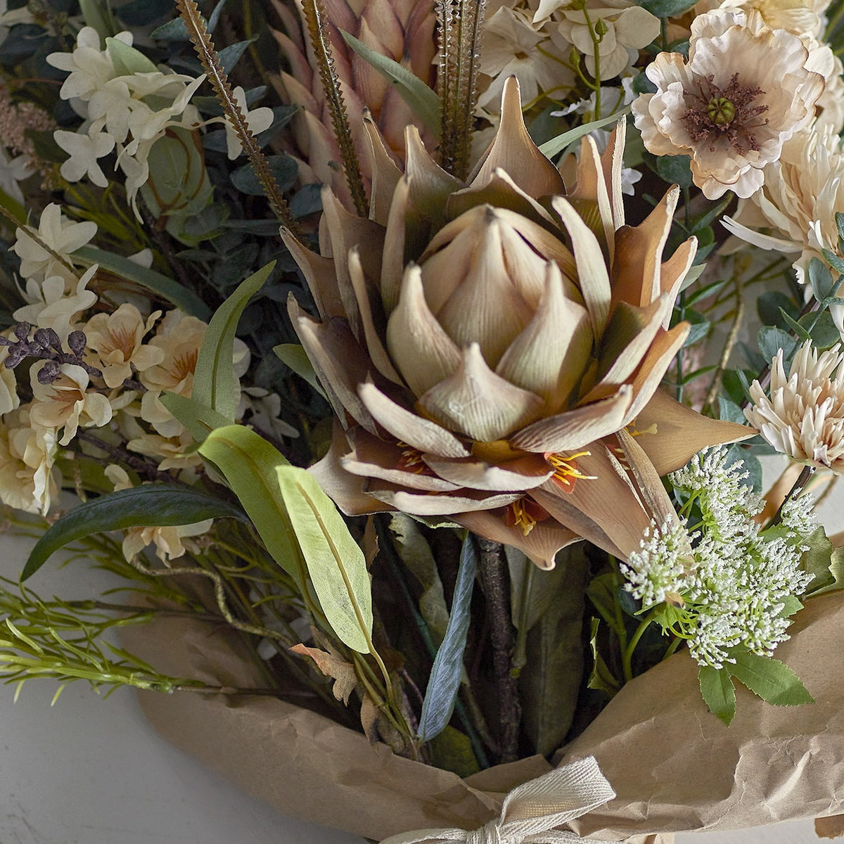 Artificial protea flower stem in beige with a large layered, pointed-petal bloom, shown in a mixed floral bouquet with greenery and wrapped kraft paper.