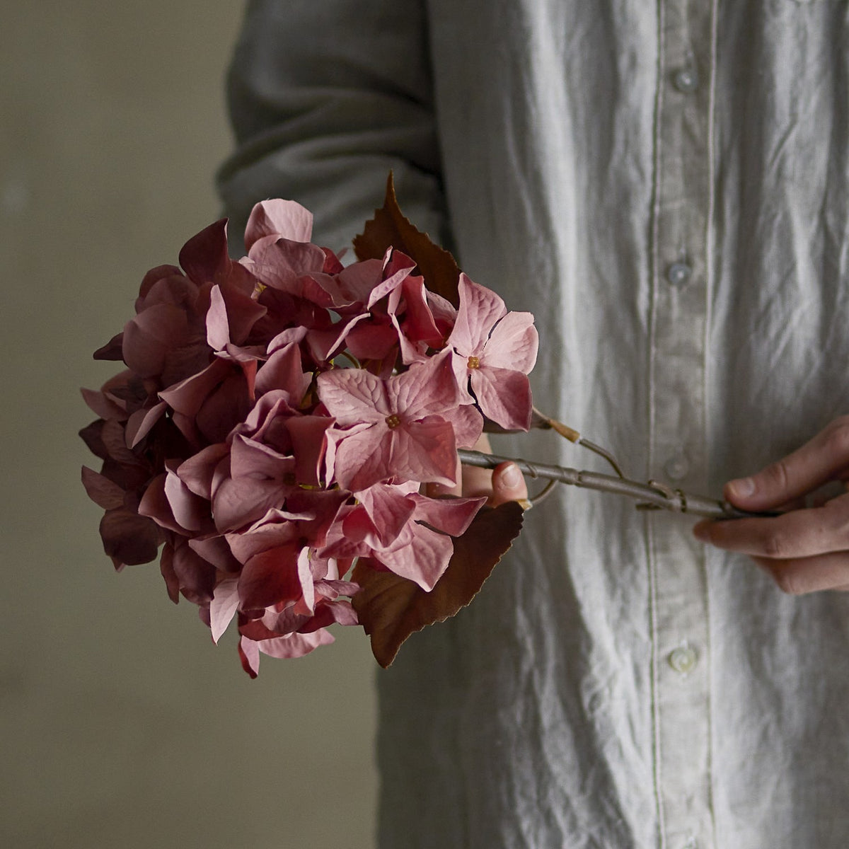 Hydrangea Artificial Stem 65cm in pink, held by hand against a person's linen shirt, showing the full rounded flowerhead and two leaves beneath it.