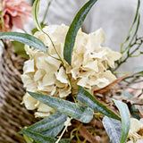 Artificial hydrangea stem in beige with a full, rounded mophead bloom, surrounded by green leaves and arranged in a woven basket display.