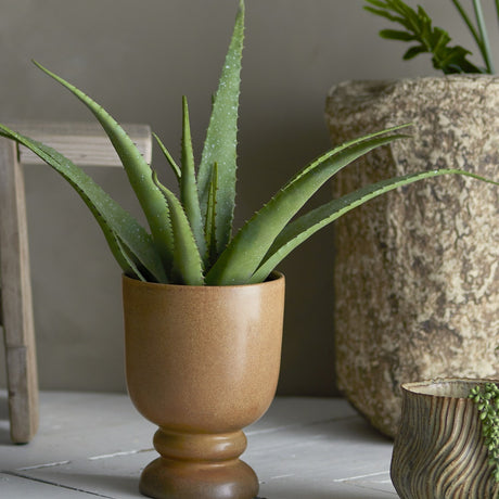 Artificial aloe plant with tall, serrated green leaves in a round tan ceramic pot, shown on a wooden floor beside stone planters.