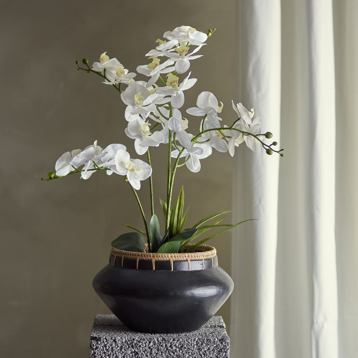 Artificial white orchid plant with arching flower stems and green leaves in a black pot with woven rim, displayed on a textured grey plinth against a neutral wall.