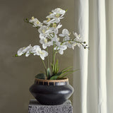 Artificial white orchid plant with arching flower stems and green leaves in a black pot with woven rim, displayed on a textured grey plinth against a neutral wall.