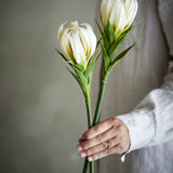 Lily Artificial Stem in white, two faux blooms with curled petals and slender green leaves, held in hand against a neutral indoor background.