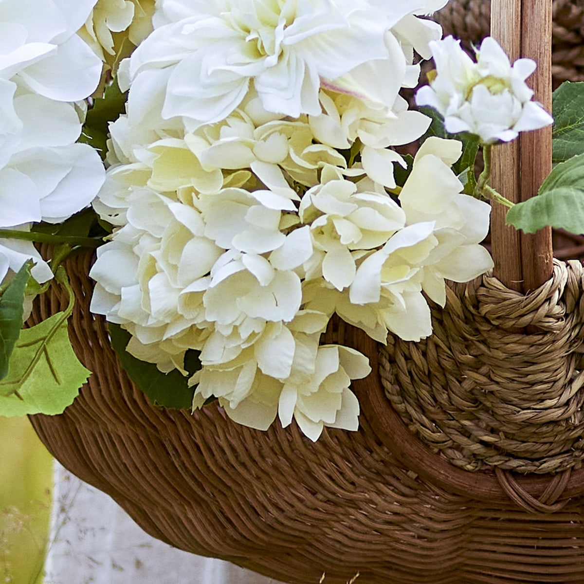 Close-up of cream artificial hydrangea stems with dense rounded florets, paired with white blooms and green leaves in a woven wicker basket.