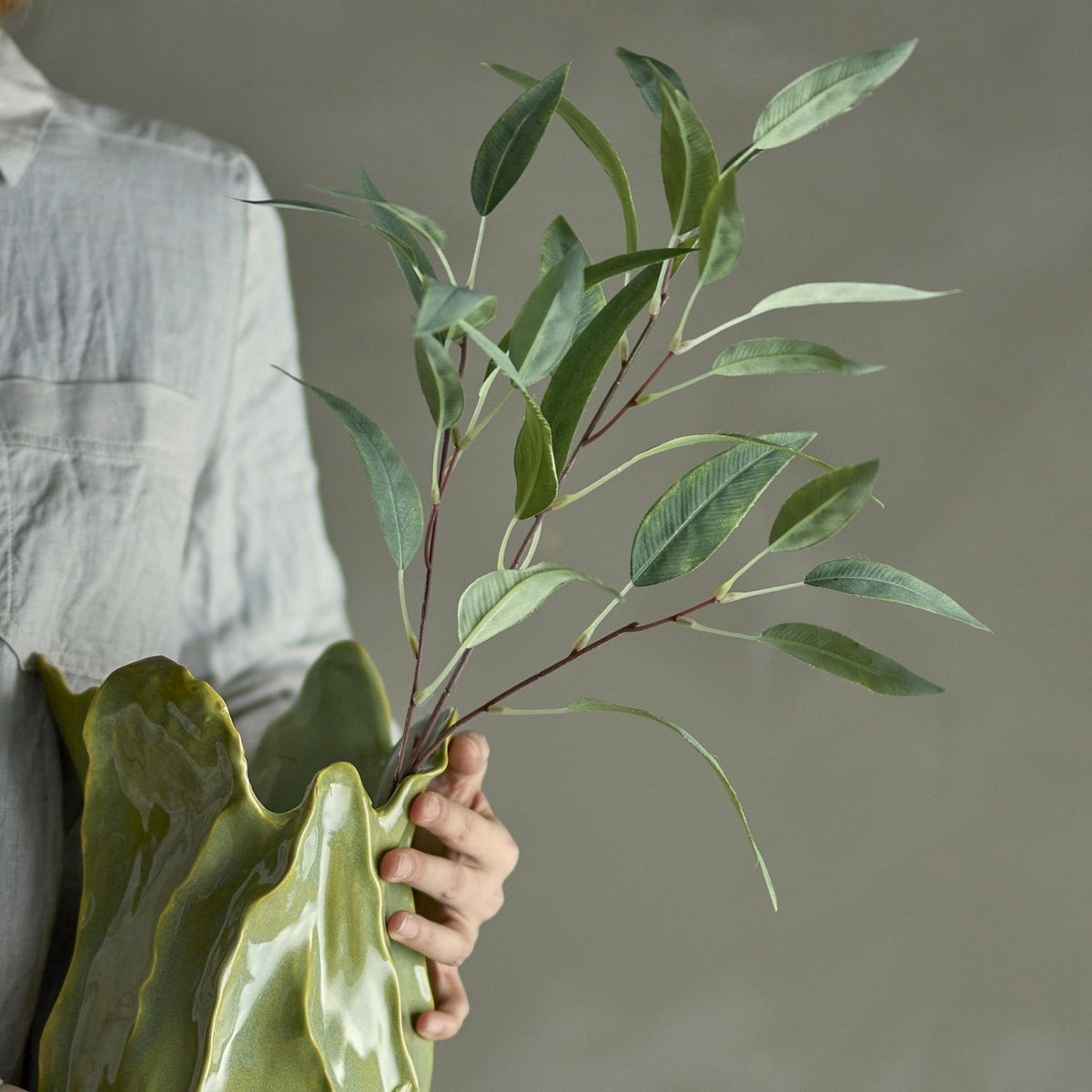 Eucalyptus Artificial Stem 85x18cm in Green held in hand against a green sculptural vase, showing slender branching stems with pointed leaves over a neutral grey background.