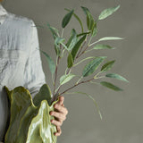 Eucalyptus Artificial Stem 85x18cm in Green held in hand against a green sculptural vase, showing slender branching stems with pointed leaves over a neutral grey background.