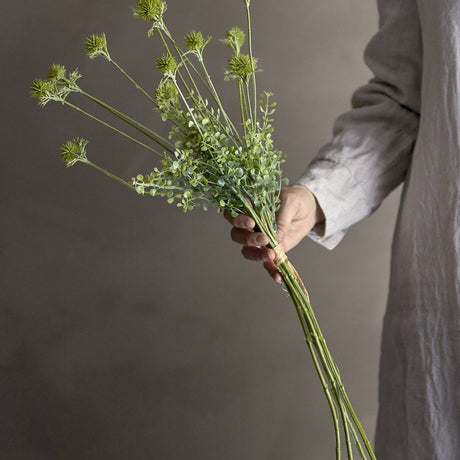 Hand holding the Wildflower Artificial Stem in green, a bunch of slender faux stems with small round spiky flowerheads and leafy sprays, tied at the base.