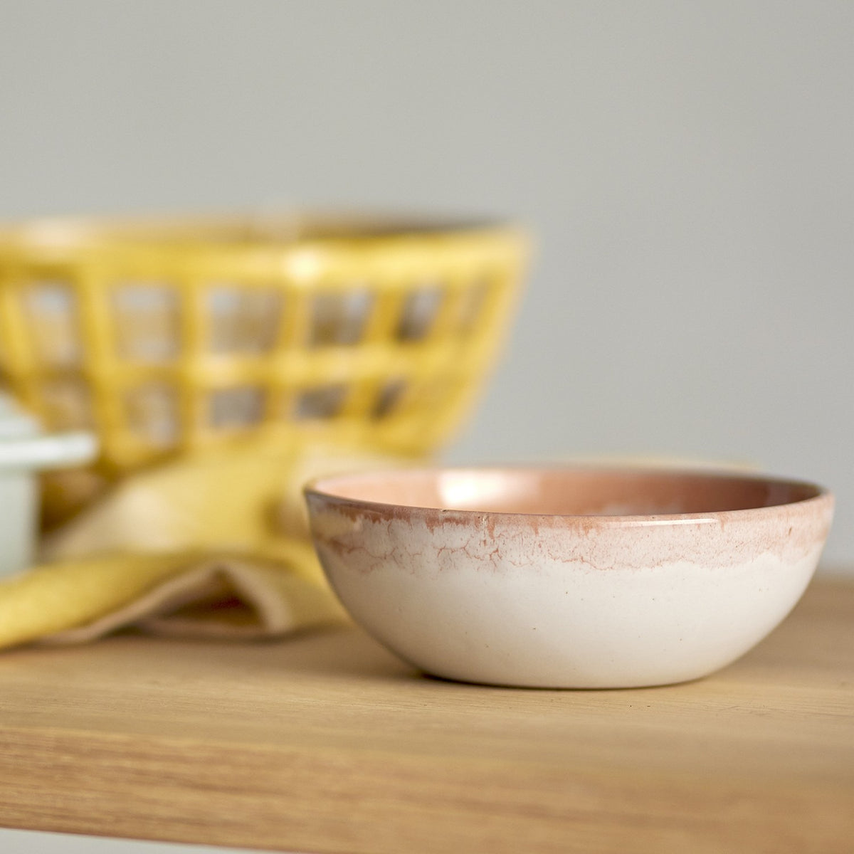 Paula Stoneware breakfast bowl from the set of 12, in orange reactive glaze with a cream base, shown on a wooden surface with a yellow woven basket blurred in the background.
