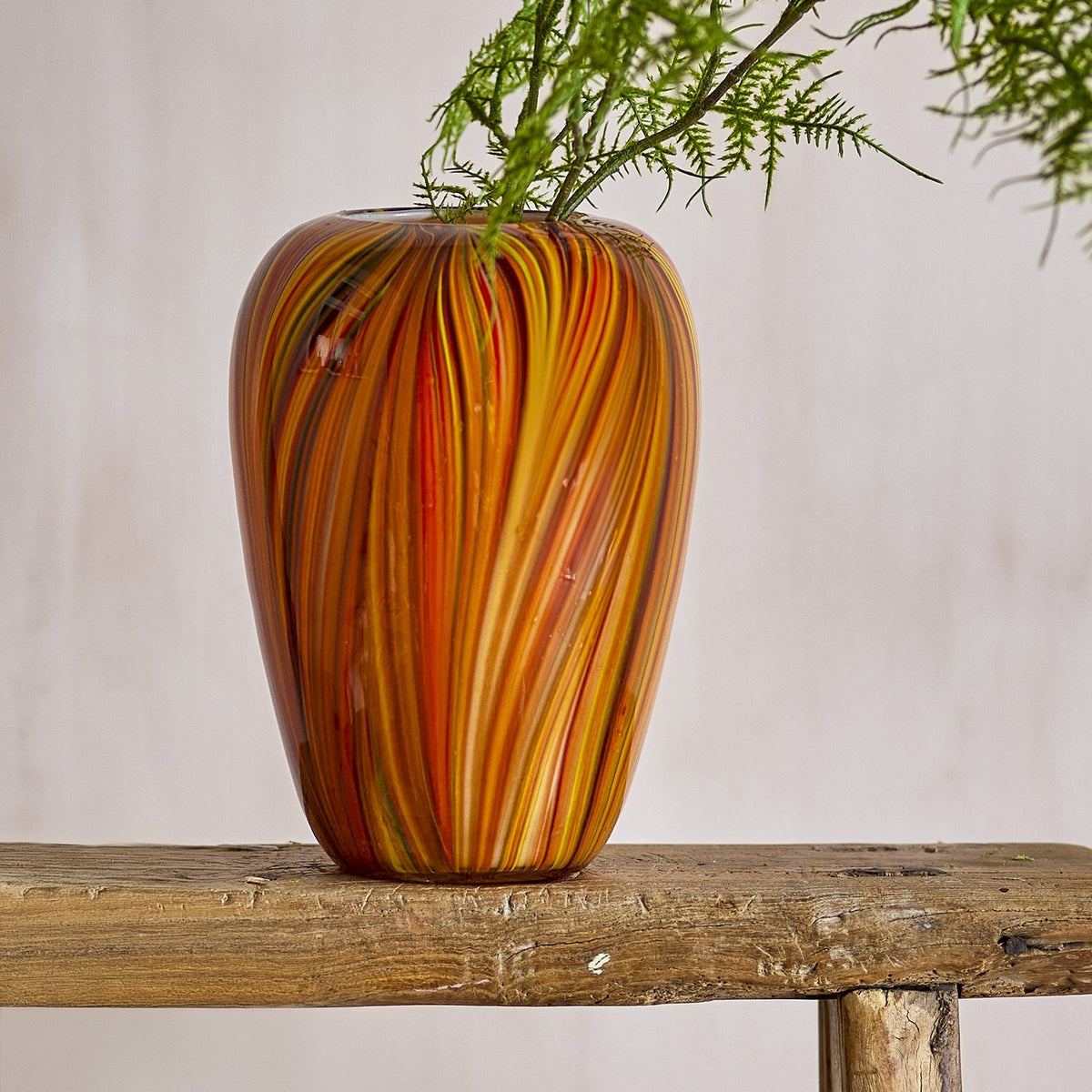 Sunny Striped Glass Vase 22.5cm in orange with fine swirling stripes and a rounded body, shown on a wooden bench with trailing greenery against a plain background.