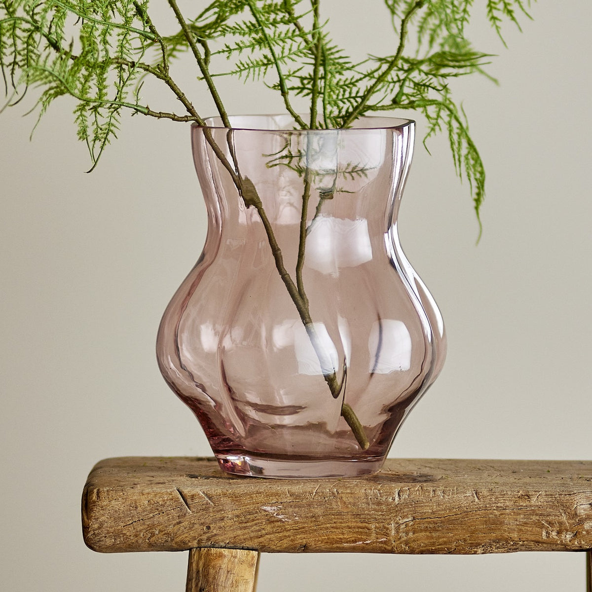 Tava Glass Vase 20cm in pink with ribbed, lobed body and flared rim, holding fern foliage on a wooden stool against a neutral background.