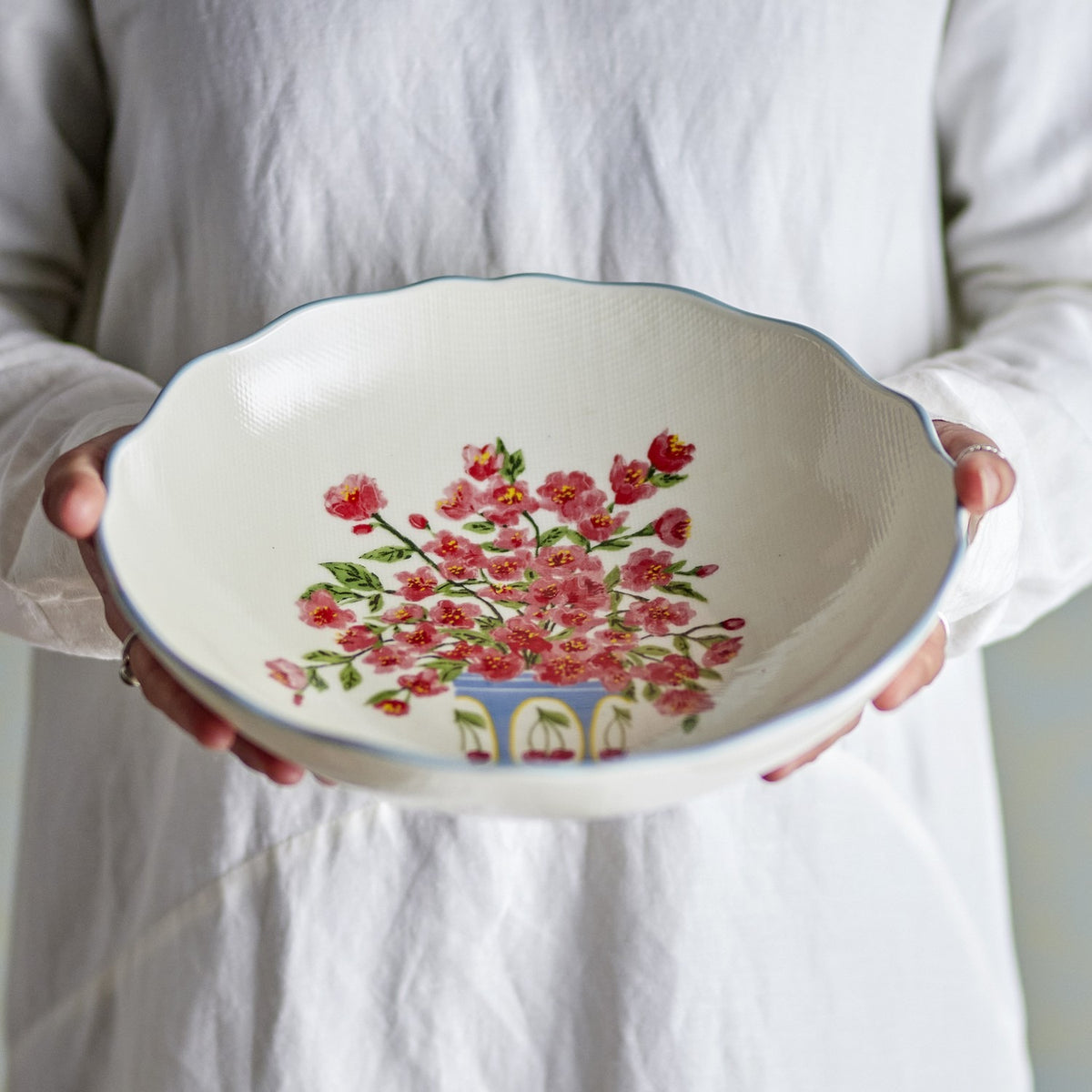 Dorothy Floral Stoneware Bowl 28cm in white, a wide shallow serving bowl with scalloped blue-edged rim and pink floral centre design, held by hands over a white outfit.