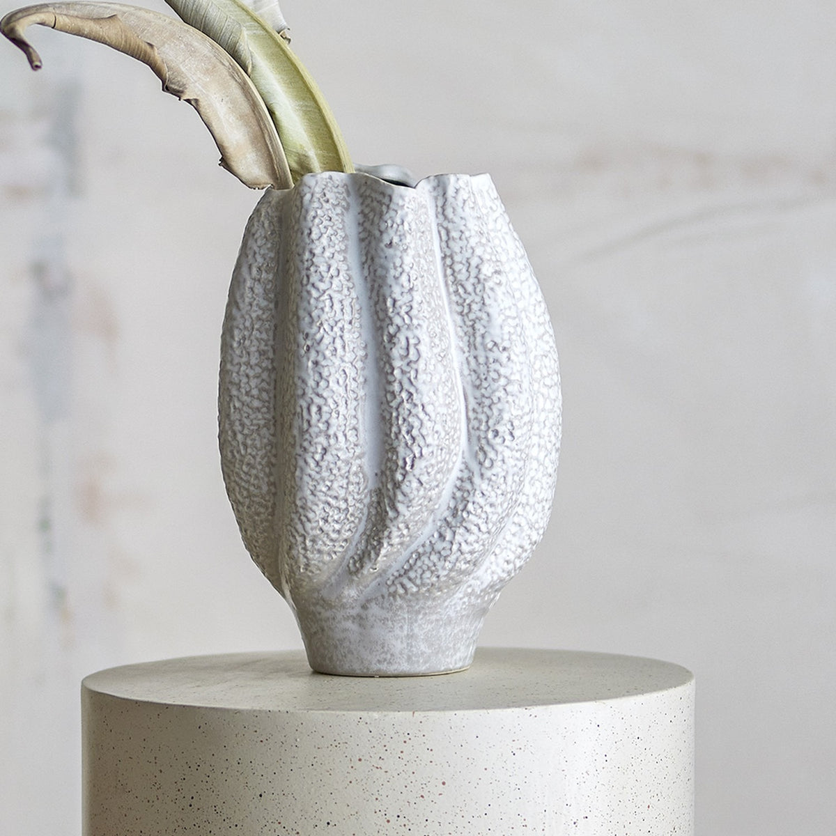 Cove Stoneware Vase 28cm in White, a ribbed ceramic vase with wavy rim and pebbled texture, shown with foliage on a concrete plinth against a neutral background.