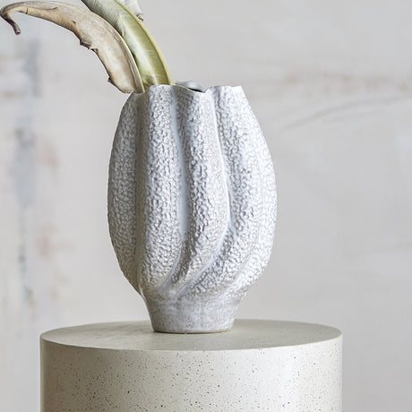 Cove Stoneware Vase 28cm in White, a ribbed ceramic vase with wavy rim and pebbled texture, shown with foliage on a concrete plinth against a neutral background.