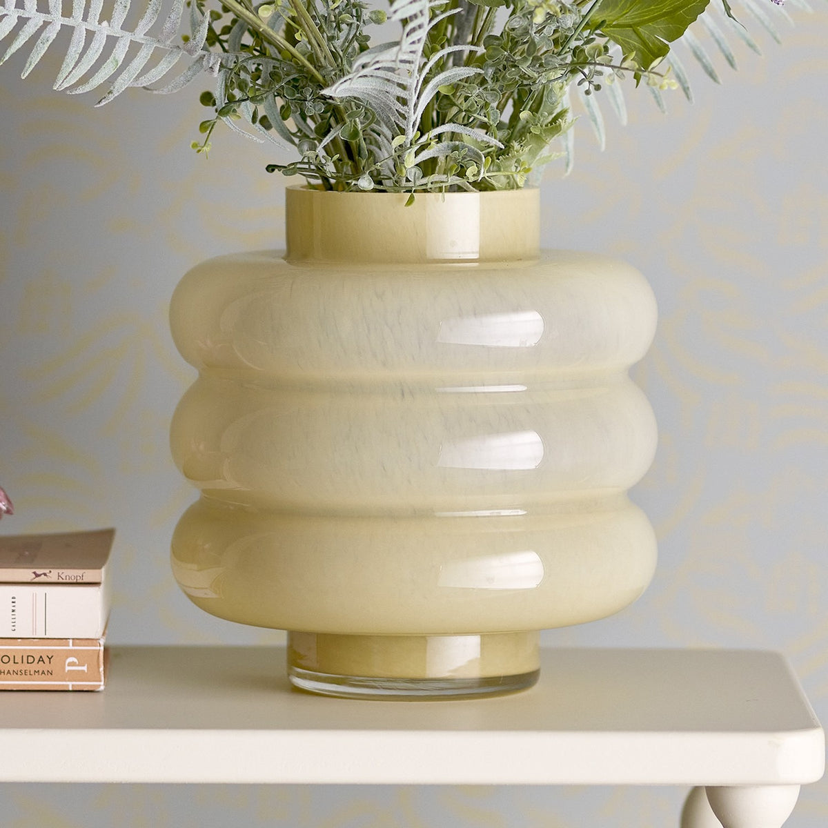 Sahara Glass Vase 22cm in Yellow, mouthblown glass with stacked rounded rings and short neck, shown on a white shelf with faux foliage against a grey patterned wall.