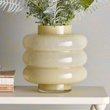 Sahara Glass Vase 22cm in Yellow, mouthblown glass with stacked rounded rings and short neck, shown on a white shelf with faux foliage against a grey patterned wall.